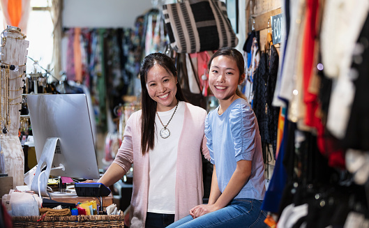 A mature Korean woman and her teenage daughter working together behind the checkout counter of a clothing store. They are smiling at the camera.