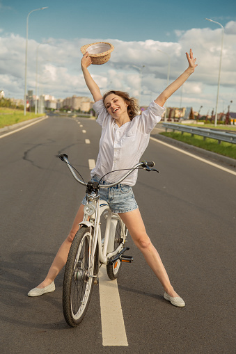 happy young woman cyclist in white blouse and hat posing with bicycle on road smiling