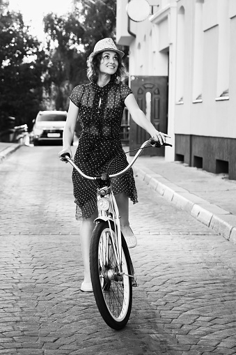 young woman in hat and dress moves on bike at old european city, sunset time, looking at camera, smiling, monochrome