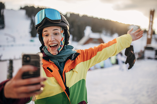 Female skier sitting sitting and using smart phone to take a selfie on a mountain ski resort.