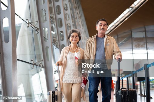 istock Cheerful senior Asian couple with wheeled luggage going for holiday vacation together 1427650042