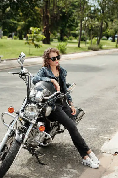 Stylish mature woman in susnglasses leaning on motorcycle when waiting boyfriend outside
