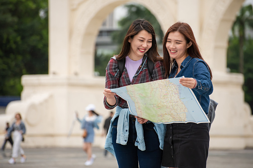 Two cheerful Asian female travelers searching direction and exploring new travel locations with tourist map in the city.