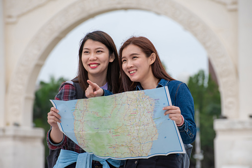 Two cheerful Asian female travelers searching direction and exploring new travel locations with tourist map in the city.