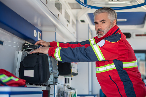Serious paramedic in a red uniform mounting a mobile ECG monitor in the medical emergency vehicle