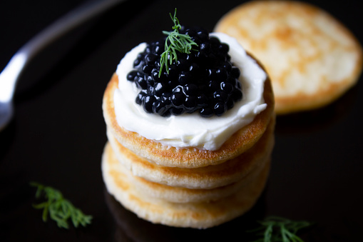 Black caviar blinis with cream cheese and fresh dill stacked. \nShot from above on a black background with a spoon and more blinis.