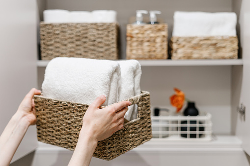 Clean fresh towels neatly folded and placed in closet organizer box. Woman putting wicker basket in bathroom wardrobe. Housework and housekeeping concepts. Selective focus on white laundry