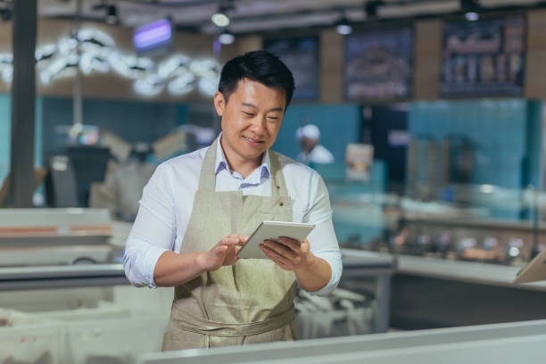 istock Asian grocery store manager salesman in apron using digital tablet counting goods in supermarket. Male worker employee doing inspection of products regular inventory to control goods online order 1427671884