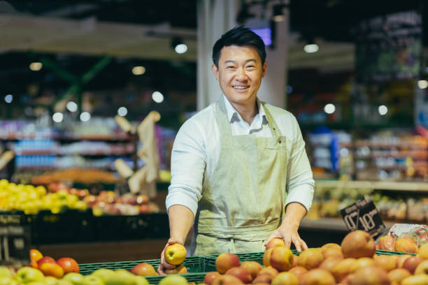 портрет продавца азиатского супермаркета, мужчины в продуктовой овощной секции, улыбающегося и смотрящего в камеру - grocer стоковые фото и изображения