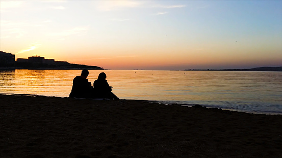 Silhouette of man and woman watching the sunset. Silhouette of the couple enjoying the sunset on the beach. Young couple sitting on a bench at sunset. There is a flying bird, silhouette