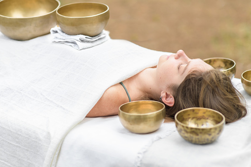 Young woman laying on a massage bed with tibetan singing bowl outdoor. Nepal copper singing bowl sound massage therapy is Buddhist healing practices. Caucasian woman relax with alternative medicine