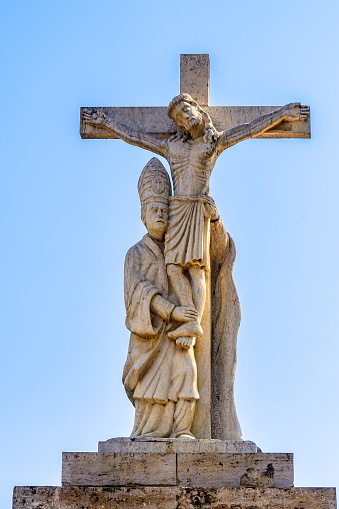 Valencia, Spain - September 29, 2022: Statue of Jesus on the Cross with a man holding his legs and waist. The statue stands on a stone slab under the blue sky.
