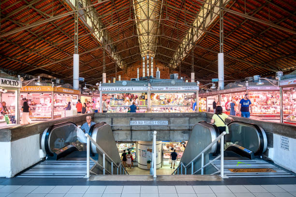 Alicante, Spain - September 29, 2022: A man and a woman on escalators inside the Mercado Central Alicante. People are shopping in the background.