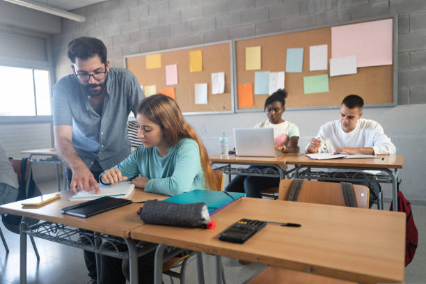 istock Young Professor helping female student in High School classroom. Friendly Teacher with beard explaining exercicse to blond girl 1427682349