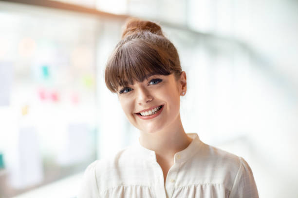 Portrait of young woman standing in modern office next to window Portrait of young woman standing in modern office next to window fringe bangs that hit at the eyebrow level stock pictures, royalty-free photos & images