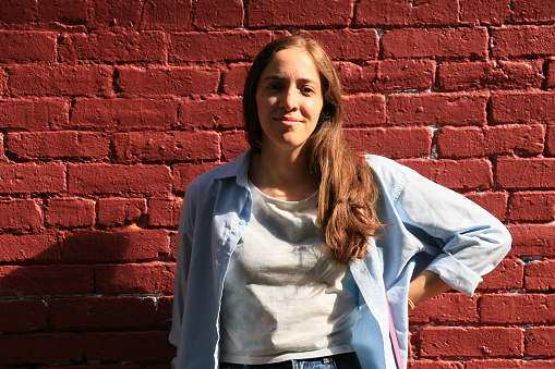 A Mexican woman standing in front of an old red brick wall. She is wearing a grey shirt and a blue denim jacket.