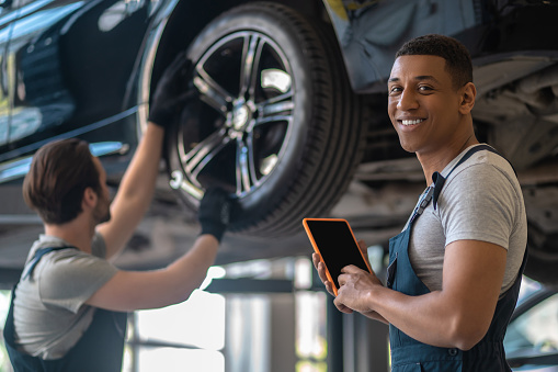 Cheerful auto repair shop worker with the tablet computer posing for the camera while fixing the car wheel