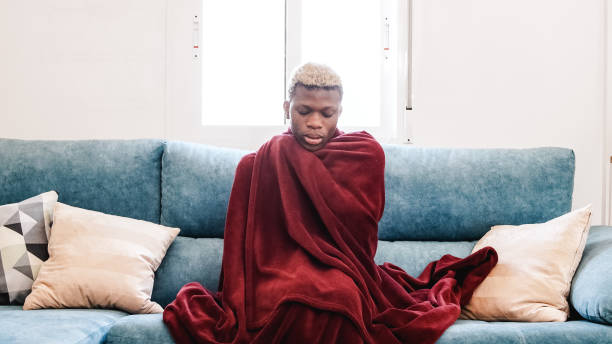 Freezing Black person wrapping in blanket on couch at home Black transgender woman with blond hair rubbing body and shaking under warm blanket while sitting on sofa in cold living room sad gay stock pictures, royalty-free photos & images
