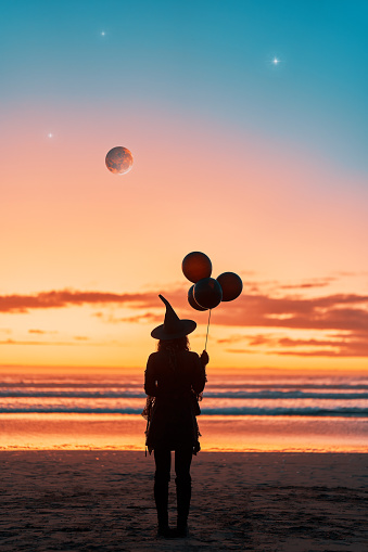 woman dressed as a witch with balloons in her hand for halloween on the beach at sunset back view