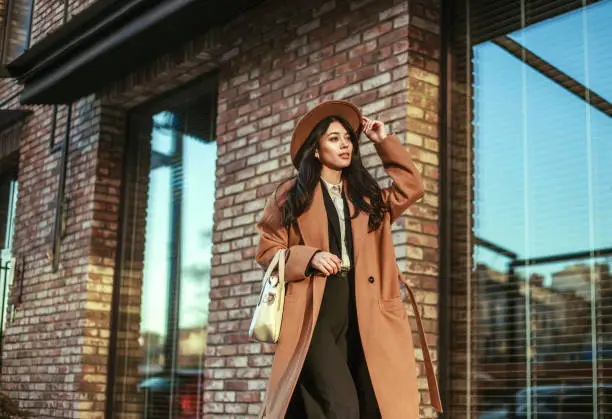 Asian business woman in a long beige coat and hat walking down the street in a loft style, on background of a brick wall, classic retro style