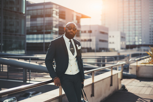 A confident mature bald bearded African man entrepreneur in sunglasses and a tailored suit is leaning against railing while standing on the street of a business district on a sunny day