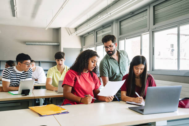 professore amichevole che spiega l'esercizio agli studenti nelle aule delle scuole superiori. giovane insegnante con la barba che aiuta la ragazza adolescente - focus sull'insegnante maschio - professor university education teacher foto e immagini stock