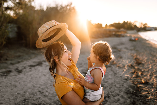 Mother holding her cute little daughter on beach in sunset, enjoying time together, playing with a pork pie hat.