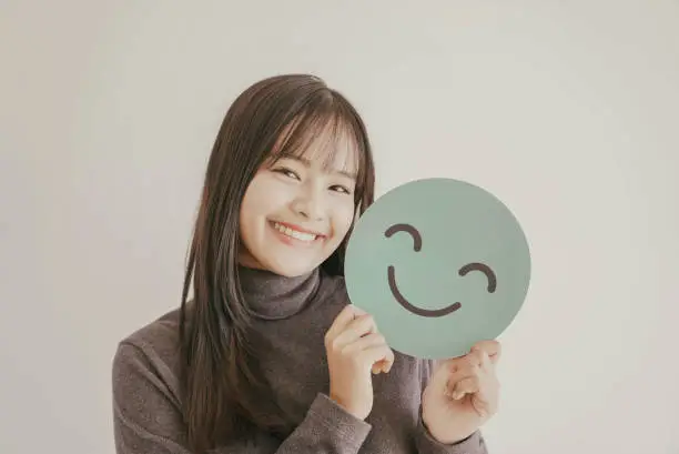 Happy young Asian woman holding smile emoji face, positive mental health concept
