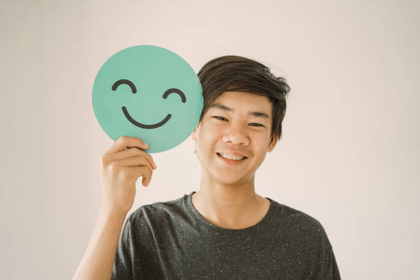 Happy mixed Asian teen boy holding smile emoji face, positive mental health concept Happy mixed Asian teen boy holding smile emoji face, positive mental health concept mental health ribbon stock pictures, royalty-free photos & images