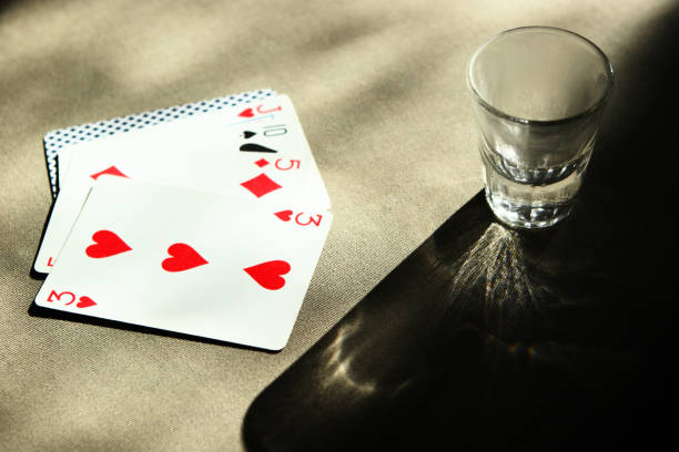 One shot glass rests on a black table surface in the upper right corner of this horizontal frame, with a hand of playing cards fanned out adjacent to it at left. \nThe card hand consists of the three of hearts atop the five of diamonds, atop the ten of spades, atop the jack of hearts, atop one card turned down.  Bright but filtered lighting enters the top of frame, casting a prism-like shadow from the shot glass onto the dark table top.  A moody, contrasty, old-style Wild West image.