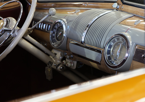 Tightly cropped horizontal image of the interior of an early 1940's vintage car, shot through the passenger side window.  Visible are parts of the steering wheel, steering column, horn, dashboard, shifter.  Dash color is a tan faux wood with gray trim.  A round speedometer and clock are prominent features on the dash.  Shot at an angle such that a small diagonal section of the amber-colored passenger side door exterior frames the bottom of the image.