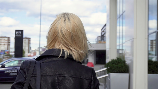 Rear view of a blonde in a black leather jacket walking in the city street. Young, blonde woman walking through the streets of the city with a bag over her shoulder.