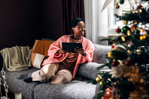 Afro-American woman comfortably sitting on the sofa in her living room, enjoying Christmas holidays, spending her time browsing social media on a tablet device.