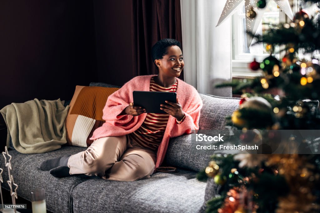 Woman Using A Tablet Device At Home Afro-American woman comfortably sitting on the sofa in her living room, enjoying Christmas holidays, spending her time browsing social media on a tablet device. Christmas Stock Photo