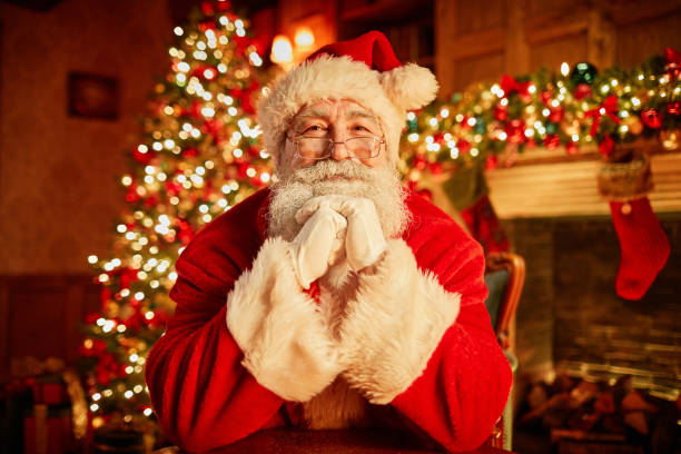 Portrait Of Traditional Santa Claus On Christmas Front view portrait of traditional Santa Claus smiling at camera resting head on arms with christmas tree in background father christmas stock pictures, royalty-free photos & images