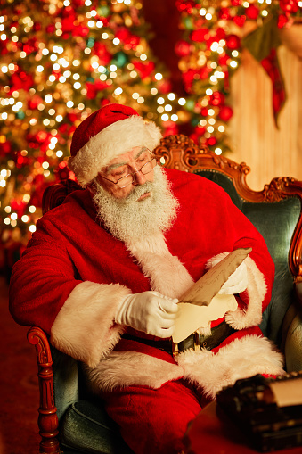 Santa Claus sits in a sofa chair in front of the fireplace in his living room, wearing pijamas with a plate of cookies and milk on his lap and a half eaten cookie in his hand, fast asleep.