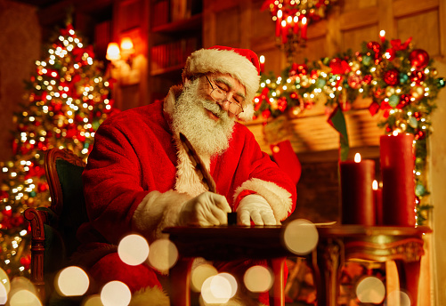 Portrait of traditional Santa Claus writing letter by fireplace on Christmas eve and smiling, copy space