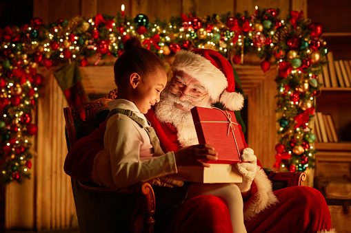 Side view portrait of cute African American girl opening Christmas present while sitting on Santas lap