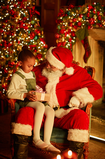 Vertical portrait of cute African American girl sitting in Santas lap and holding Christmas present in cozy setting