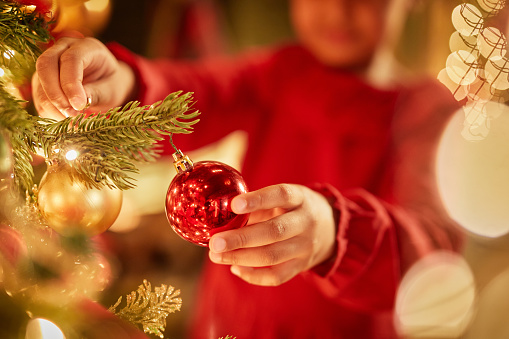 Closeup of unrecognizable little girl decorating Christmas tree with ornaments and twinkling lights, copy space