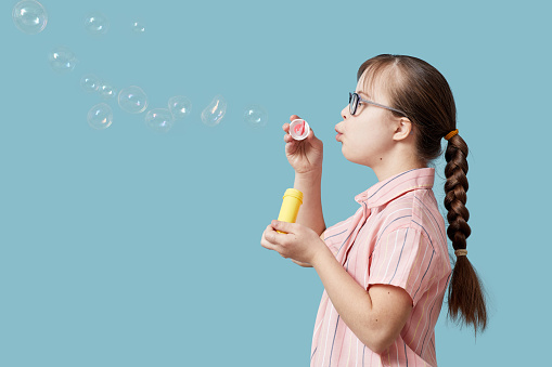 Side view portrait pf playful girl with Down syndrome blowing bubbles against blue background