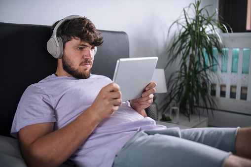 Man with wireless headphones at home concentrated on winning the mobile game that he plays on digital tablet.