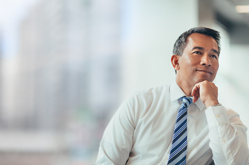 Portrait of Asian mature businessman with hand on chin sitting thinking satisfaction looking forward away in bright business office in businesswear