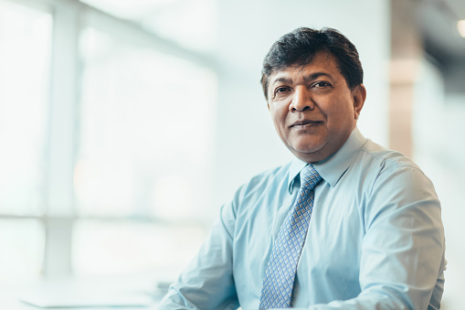 Portrait of Asian Indian mature businessman sitting in bright office in businesswear