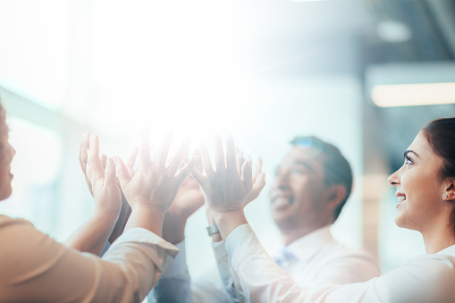 Multiracial group of business persons students clasping hands in stacked hands high-five lifting concept in team building success educational classroom office looking up with bright light