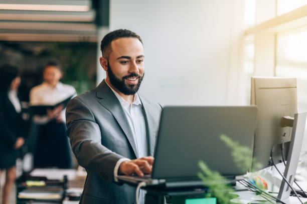 Multiracial Portuguese Jamaican mid adult businessman with beard standing smiling at desk with laptop reviewing data in bright business office wearing suit stock photo