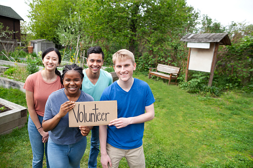 Multiracial group of young men and young women stand together and hold volunteer sign within community garden park in neighborhood