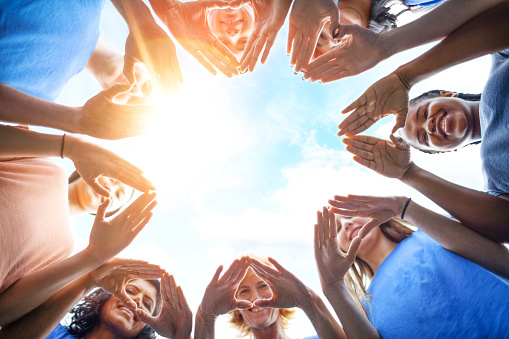 Multiracial group of diverse people stand in circle as community volunteers to show support and commitment to teamwork success togetherness making hand gesture in concept symbol sign of heart shape outdoors with sky