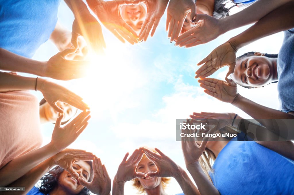 Multiracial group of diverse people stand in circle as community volunteers to show support and commitment to teamwork success togetherness making hand gesture in concept symbol sign of heart shape outdoors with sky Community Outreach Stock Photo