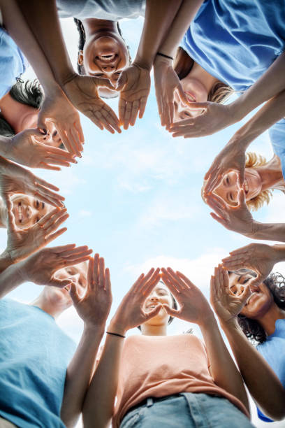 multiracial group of diverse people stand in circle as community volunteers to show support and commitment to teamwork success togetherness making hand gesture in concept symbol sign of heart shape outdoors with sky - youth organization imagens e fotografias de stock